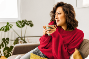 A woman sitting on her couch in a red sweater holding a mug, and looking straight ahead, with a relaxed facial expression