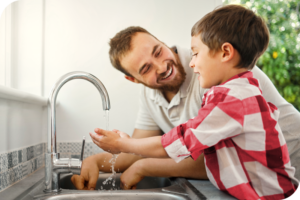 A man and a child smile as they both wash their hands in a kitchen sink