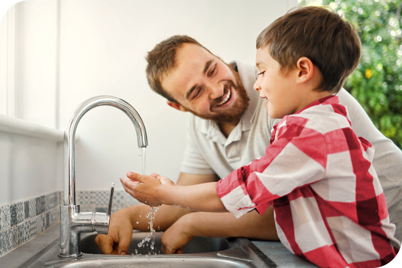A man and a child smile as they both wash their hands in a kitchen sink