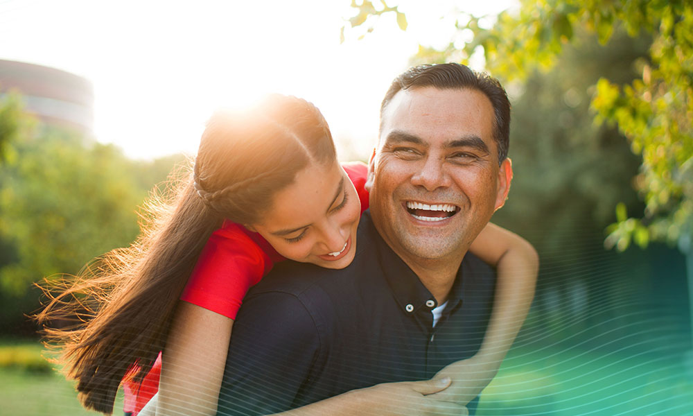 A playful latin mature father carrying his teen daughter on the back, looking away and laughing in a horizontal waist up shot outdoors with backlight.