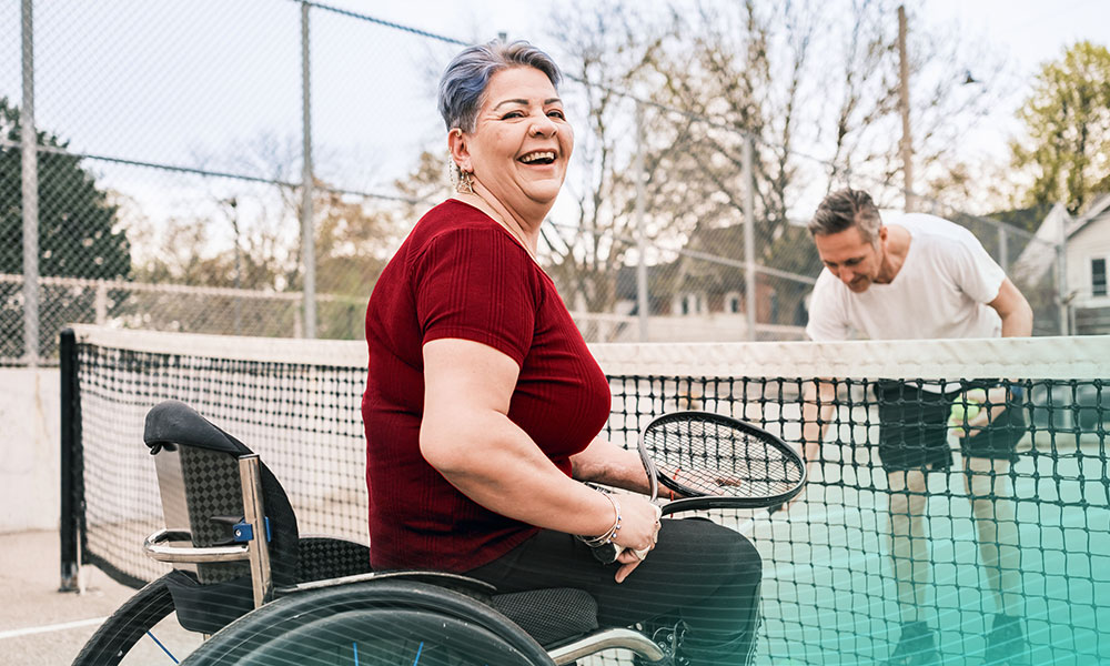 Disabled Latin woman practice wheelchair tennis with her coach outdoors. She has both artificial legs and disfigured left palm. Both dressed in casual sport clothes. Exterior of public tennis court in the public city park during the day.