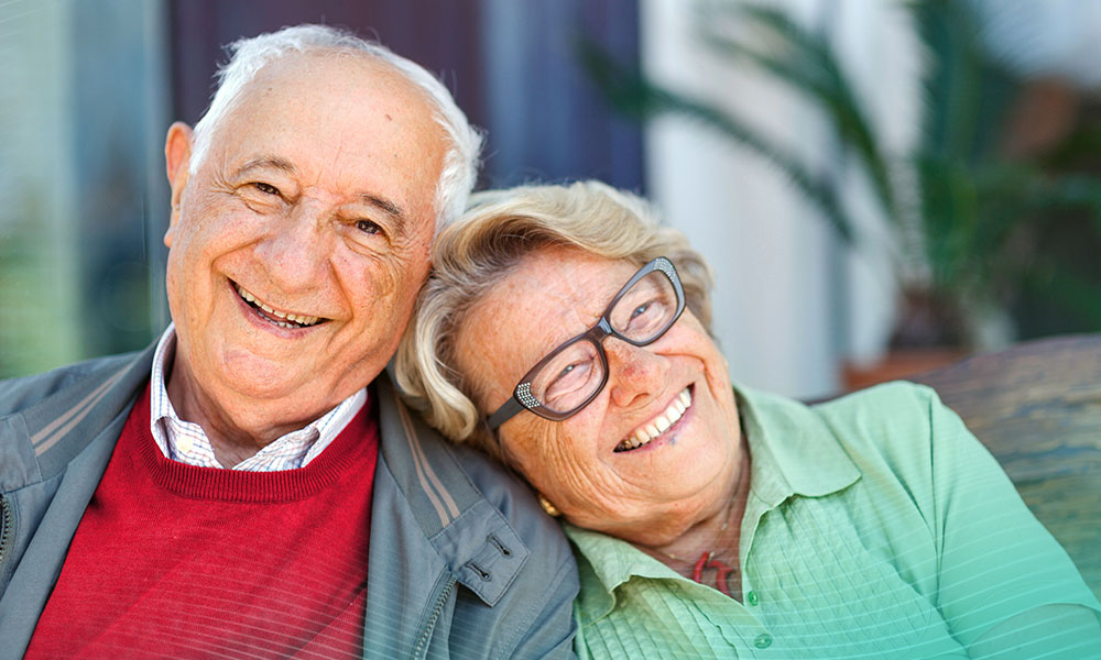 A senior couple is laughing. She has her head on his shoulder. She is wearing glasses and a green shirt. At her neck, she has a red pendant. He is wearing a blue jacket, a brown sweater and a white shirt. They are sitting on a wooden bench.