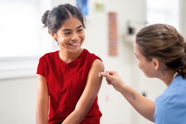 A young Hispanic girl sits up on an exam table in the doctors office as she receives a vaccination. She is dressed casually and the nurse is seated beside her in blue scrubs as she places a bandage over the injection site.